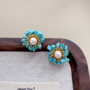 May include: A pair of turquoise and gold-tone stud earrings. Each earring features a cluster of small, irregular turquoise stones arranged around a central white pearl. The earrings are set on a gold-tone metal base.