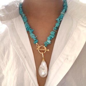 May include: A necklace featuring turquoise stone chips, a gold-coloured clasp, and a large, white, pear-shaped pearl pendant. The necklace is worn over a white shirt.