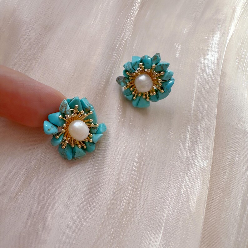 May include: Flower-shaped earrings with turquoise stone petals and a central pearl. The earrings have a gold-colored setting. The design is floral and the earrings are set against a soft, off-white background.