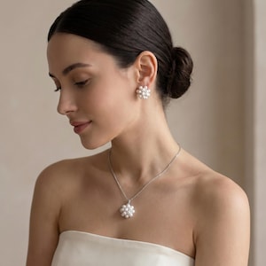 May include: A jewellery set consisting of a necklace and matching stud earrings. Each piece features a cluster of small, round, white pearls arranged in a circular floral pattern, suspended on a thin, silver-toned metal chain.