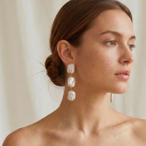 Peut inclure: Boucles d'oreilles pendantes dorées composées de trois pierres rectangulaires blanches crémeuses. Chaque pierre est sertie dans un cadre doré, créant un design simple et élégant.