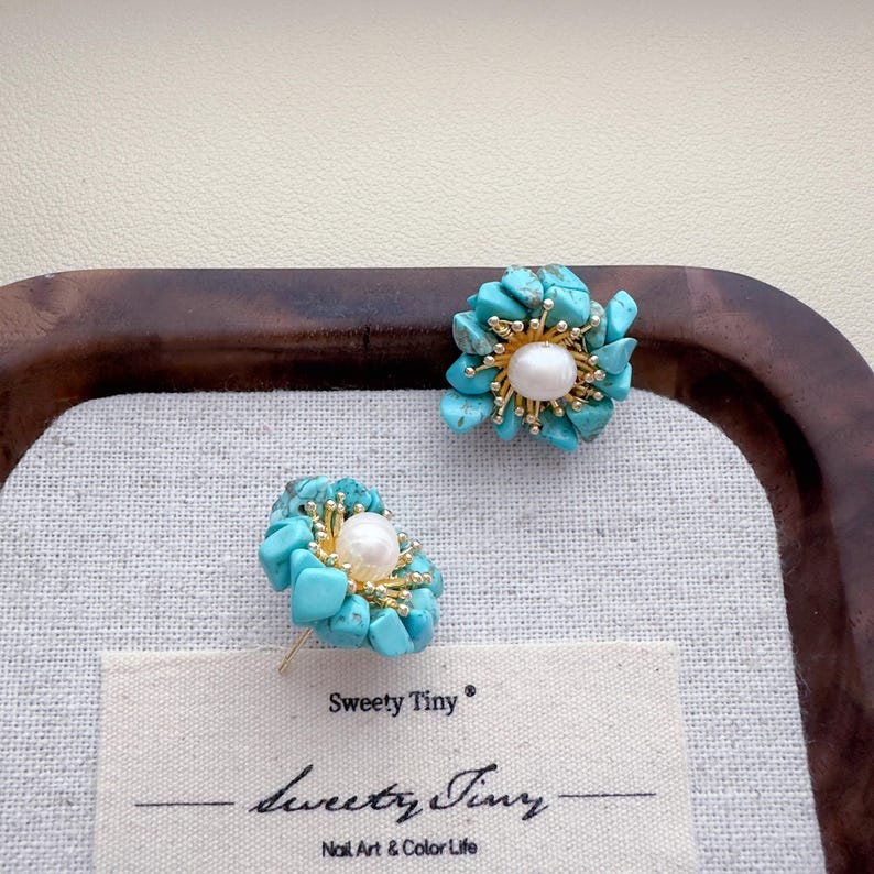 May include: A pair of flower-shaped stud earrings. Each earring features a cluster of turquoise stones surrounding a central white pearl. The earrings have gold-colored accents and are displayed on a white surface with the text "Sweety Tiny".