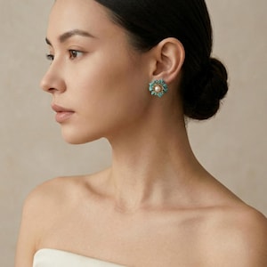 May include: A close-up of a person wearing a turquoise and pearl flower-shaped earring. The earring has a central pearl surrounded by turquoise stones, creating a floral design. The person's skin tone is fair, and their hair is styled in a bun.