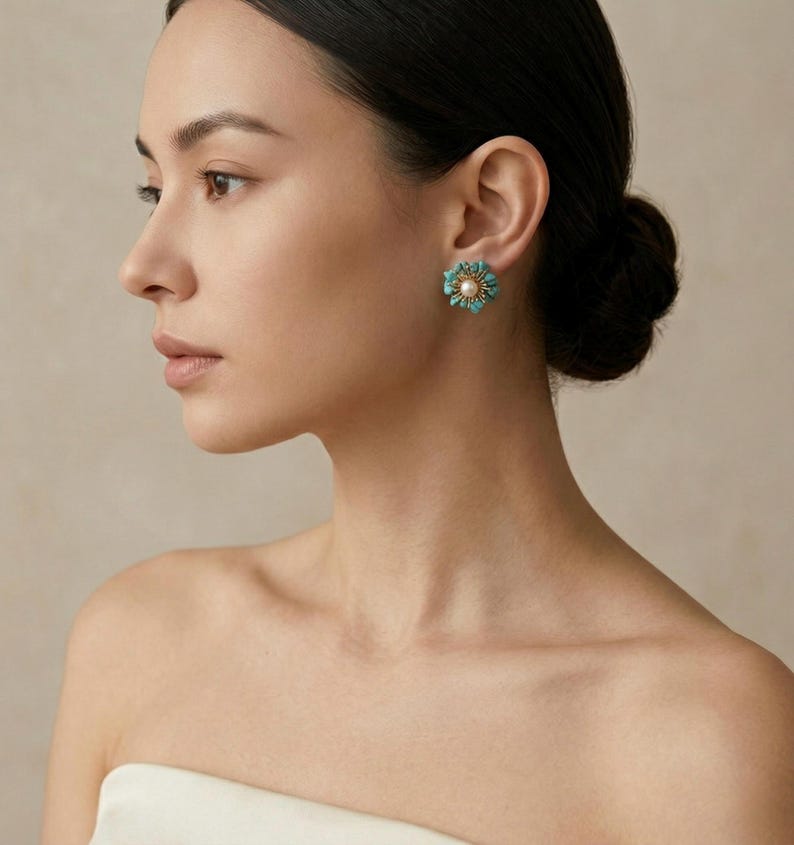 May include: A close-up of a person wearing a turquoise and pearl flower-shaped earring. The earring has a central pearl surrounded by turquoise stones, creating a floral design. The person's skin tone is fair, and their hair is styled in a bun.