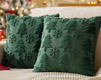 Può includere: Due cuscini decorativi verdi con un motivo a forma di albero di Natale. I cuscini sono quadrati e hanno una superficie strutturata. Lo sfondo mostra un albero di Natale con luci.