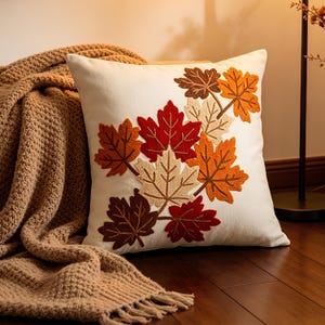 May include: A square white throw pillow featuring an embroidered cluster of maple leaves in shades of red, orange, brown, and cream. The leaves are arranged in a diagonal pattern across the front surface, showcasing detailed stitching and textures.