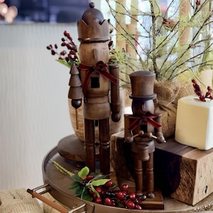 May include: Two dark brown wooden nutcracker figures. The taller one wears a crown and holds a small tree. The shorter one wears a top hat and holds drumsticks over a drum. Both figures feature dark red velvet ribbon bows at their necks.