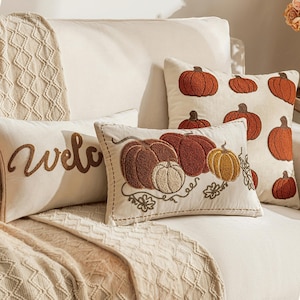 Pode incluir: Três almofadas retangulares e quadradas de tecido cor de creme. Uma almofada apresenta o texto bordado em castanho "Welcome". Duas almofadas exibem desenhos de abóboras texturizados e em relevo em tons de laranja, castanho e bege com vinhas verdes.