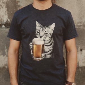 以下が含まれることがあります： ビールジョッキを持った猫の白黒イラストがプリントされた濃いグレーのTシャツ。猫は真剣な表情をしています。ビールジョッキには黄金色のビールと白い泡が入っています。