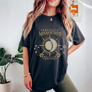 Op de afbeelding: Donkergrijs t-shirt met de tekst "PERFECTLY MISALIGNED" boven een gouden en witte zon-maan afbeelding met bloemendetails. Het t-shirt heeft een relaxte pasvorm.