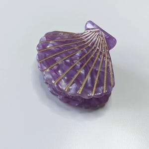 Könnte beinhalten: Eine muschelförmige Haarspange in einem durchscheinenden Violett mit marmorierter Textur. Die Oberfläche weist erhabene, goldfarbene metallische Rippen auf, die von der Scharnierseite ausgehen und die Struktur einer Muschel nachahmen.