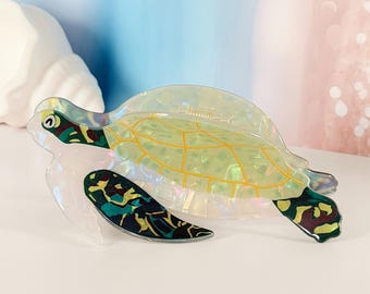 May include: A sea turtle shaped hair claw clip made of translucent iridescent material. The shell features a yellow grid pattern, while the head and flippers are decorated with dark green, black, and brown camouflage-like patches and a small eye.