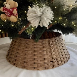 Puede incluir: Un collar para árbol de Navidad de mimbre trenzado en color marrón claro con forma cilíndrica cónica. El collar se sitúa en la base de un árbol de Navidad artificial verde decorado con adornos florales blancos y un pequeño oso de peluche con un suéter rojo.