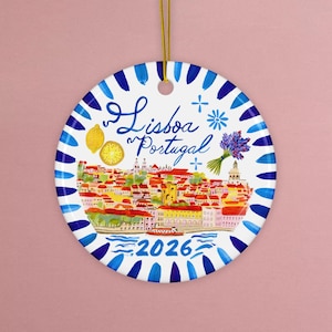 May include: A round ceramic ornament with a watercolor illustration of Lisbon, Portugal. The ornament features a blue and white border, the text "Lisboa Portugal" and the year "2026". A gold string is attached for hanging.
