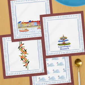 May include: A collection of white square napkins with various designs. One features a cityscape with a sailboat, another a carousel with "Copenhagen Denmark" text, a third with orange flowers, and the last with swan illustrations. Each napkin has a blue border.