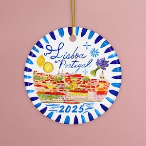 May include: A round, white ceramic ornament with a blue and white striped border. The ornament features a watercolor illustration of Lisbon, Portugal, with the text "Lisboa Portugal" and the year "2025". A gold string is attached for hanging.