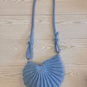 Puede incluir: Bolso de crochet hecho a mano, azul claro, con una correa larga. El bolso tiene un diseño único en forma de abanico con un detalle circular central. La correa está sujeta con anillos dorados. El bolso está hecho de hilo suave y texturizado.