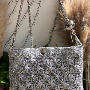Op de afbeelding: Een zilveren gehaakte handtas met een bloemenpatroon. De tas heeft een zilveren kettingriem en een magnetische sluiting. De tas is ongeveer 20 cm breed en 15 cm hoog.
