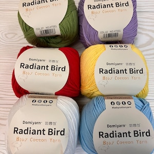 Könnte beinhalten: Sechs Knäuel Domiyarn Radiant Bird Baby Cotton Yarn in verschiedenen Farben: Grün, Lila, Rot, Gelb, Weiß und Blau. Jedes Knäuel hat ein Etikett mit dem Markennamen und der Garnart.