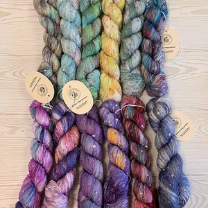 Op de afbeelding: Een verzameling kleurrijke garen strengen in verschillende tinten blauw, groen, geel, paars en rood. Elke streng is versierd met pailletten en heeft een label. Het garen is gerangschikt op een lichtgekleurd houten oppervlak.