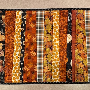 Puede incluir: Un camino de mesa acolchado con temática otoñal. Presenta varias telas con diferentes patrones, incluyendo calabazas, hojas y cuadros. El camino de mesa está bordeado con un ribete negro.