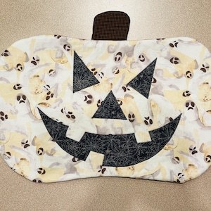 May include: A white pumpkin-shaped fabric pot holder with a black jack-o'-lantern face. The fabric is printed with a pattern of white ghosts on a beige background.