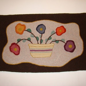 May include: A brown rug with a white center featuring a hand-stitched design of five colorful flowers in a cream-colored pot with purple, blue, and white stripes.