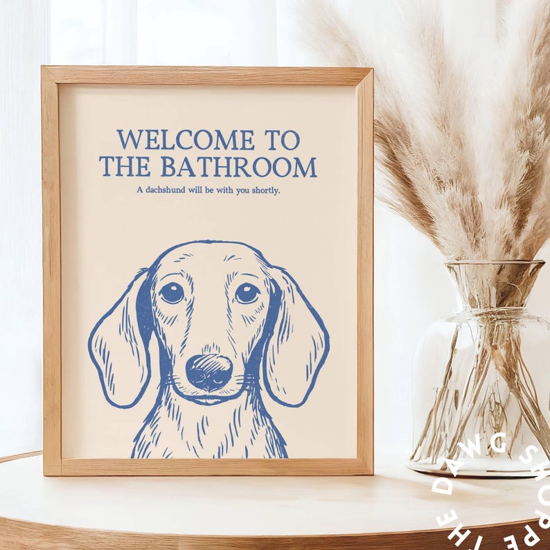 Peut inclure: Un cadre en bois clair contenant une illustration de teckel bleu sur fond cr&egrave;me. Le texte "WELCOME TO THE BATHROOM" est au-dessus du chien, avec "A dachshund will be with you shortly." en dessous.