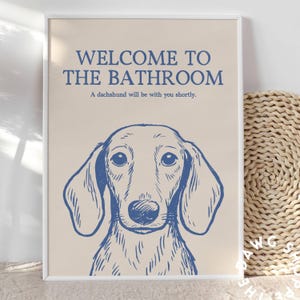 Peut inclure: Une affiche encadr&eacute;e avec une illustration de teckel bleu sur fond beige. Le texte dit "WELCOME TO THE BATHROOM" au-dessus de "A dachshund will be with you shortly."