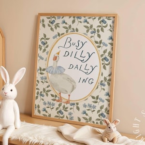 May include: A framed art print featuring a whimsical illustration of a goose wearing a party hat and a ruffled collar, with the words "Busy Dilly Dally Ing". The background has a floral pattern. A white stuffed rabbit sits nearby.