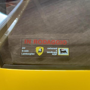 Puede incluir: Una ventana de coche amarilla con una pegatina roja que dice "IN RODAGGIO". Debajo, logotipos de Lamborghini y AGIP, con texto en italiano. El interior del coche es visible.