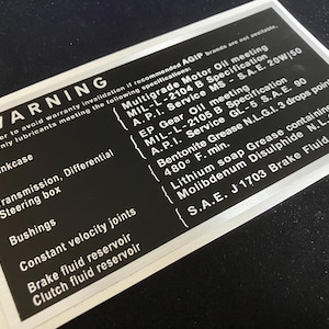 May include: A black and white warning label with silver trim. The label has the word "WARNING" in large white letters at the top. The label lists different vehicle parts and the types of fluids to use for each part. The label also includes a list of fluid specifications, including MIL-L-2104 B, A.P.I. Service MS, S.A.E. 20W/50, MIL-L-2105 B, A.P.I. Service GL-5, S.A.E. 90, Bentonite Grease N.L.G.I. 3 drops point, 480° F. min., Lithium soap Grease containing 5%, Molibdenum Disulphide N.L.G.I. 2, and S.A.E. J 1703 Brake Fluid.