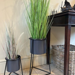 May include: Two grey metal planters with black geometric stands. One contains tall green grass, the other a mix of dried grasses. The planters are on a tiled floor, suitable for indoor decoration.