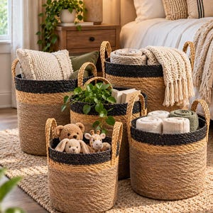 May include: A set of four cylindrical woven baskets featuring natural tan fibre bodies with black woven rims and two side handles. The baskets contain various items including textured pillows, a green plant, plush toys, and folded fabric blankets.
