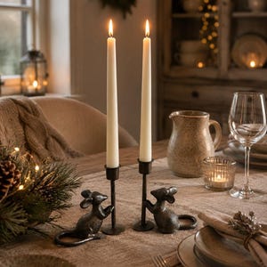 May include: Two dark metal candle holders, each featuring a sculpted mouse figure at the base holding a tall, slender, off-white taper candle. The candles are lit, emitting a warm glow, and the holders are placed on a textured linen table runner.