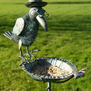 May include: A decorative metal bird feeder in the shape of a bird wearing a top hat and spectacles. The bird is perched above a leaf-shaped bowl filled with birdseed. The sculpture is painted in blue and green, set against a green lawn.
