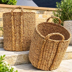May include: Two cylindrical woven baskets made of natural tan seagrass. Each basket features a tight, textured weave and two sturdy, braided vertical handles that extend from the rim down the sides, providing a structured and durable design.