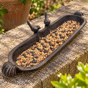May include: An elongated, dark-coloured metal bird feeder tray with a textured finish. The tray features two small, sculpted bird figures perched on one side and decorative shell-like motifs at each end, filled with a mix of small seeds.