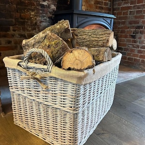 May include: A grey wicker log basket filled with firewood, positioned in front of a fireplace. The basket features a burlap lining and rope handles. The logs vary in size and shape, with visible bark and moss.