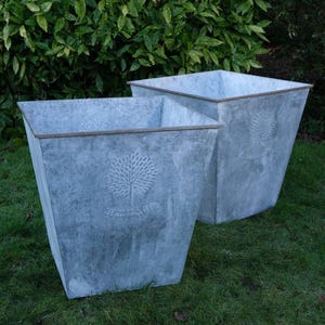 May include: Two galvanised metal planters with a square shape and a slightly tapered design. Each planter features an embossed tree design and the word "HERITAGE". The planters are set on a green lawn.