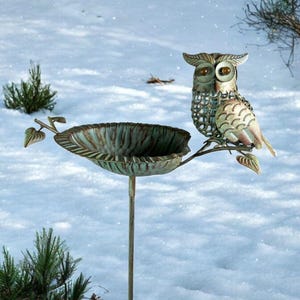May include: A metal owl bird feeder with a distressed finish. The owl sits on a branch above a leaf-shaped bowl. The feeder is set against a snowy backdrop, with evergreen shrubs in the foreground.