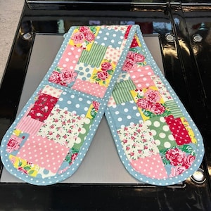 May include: A patchwork oven glove with a colourful design. It features squares of pink polka dots, floral patterns, and green stripes, set against a light blue polka dot background with a light blue trim.