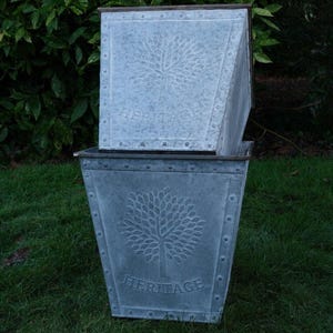May include: Two galvanized metal planters with a tree design and the word "HERITAGE" embossed on the front. The planters are square with a slightly tapered shape and are riveted along the edges. They are sitting on green grass.