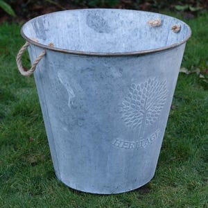 May include: A grey galvanised metal bucket with a tree design and the word "HERITAGE" embossed on the side. It has rope handles and sits on green grass. The bucket is a light grey colour.