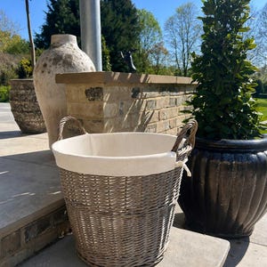 May include: A round, grey woven wicker basket featuring two sturdy handles on opposite sides. The interior is lined with a smooth, off-white fabric that folds over the top rim, secured with small fabric ties on the exterior of the basket.
