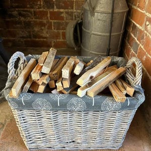 May include: A grey wicker log basket filled with firewood. The basket has rope handles and a grey fabric lining with a white tree pattern. The firewood is a mix of light and dark wood pieces, ready for a fireplace.