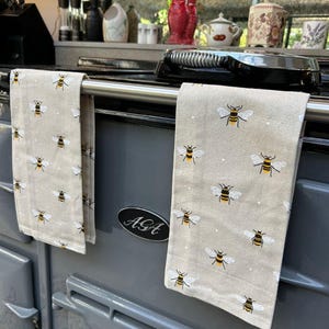 May include: Two beige kitchen towels featuring a repeating pattern of yellow and black bees with white wings, draped over the silver handle of a grey AGA stove. The stove has a black oval emblem with the white text AGA centred on the front panel.