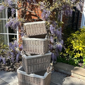 May include: A stack of four rectangular woven wicker baskets in a natural light brown finish. Each basket features a removable off-white fabric liner tucked inside the rim. The baskets are arranged in a vertical column, decreasing in size from bottom to top, showcasing a consistent weave pattern and sturdy construction throughout the set.