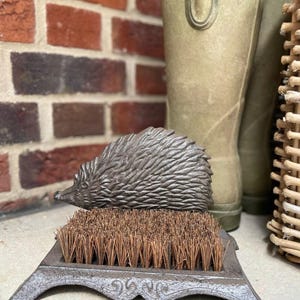 May include: A cast iron boot scraper with a hedgehog design. The scraper has a bristled brush and a decorative base. The hedgehog sits above the brush. The color is a dark, weathered grey. The scraper is likely used to clean muddy boots.