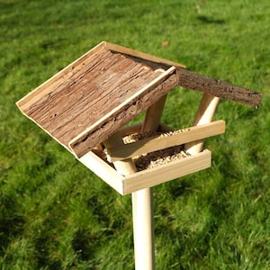 May include: A wooden bird feeder with a natural bark roof and a rectangular feeding tray. The feeder is mounted on a light-coloured wooden pole and is set against a backdrop of green grass. The feeder is filled with birdseed.